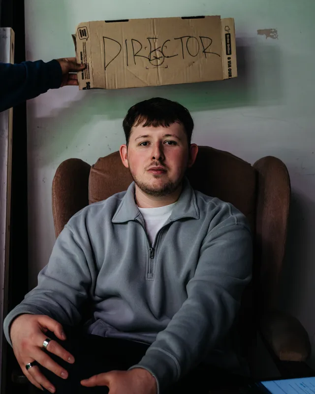 Kian sitting in a worn armchair under a hand-written cardboard sign that reads Director